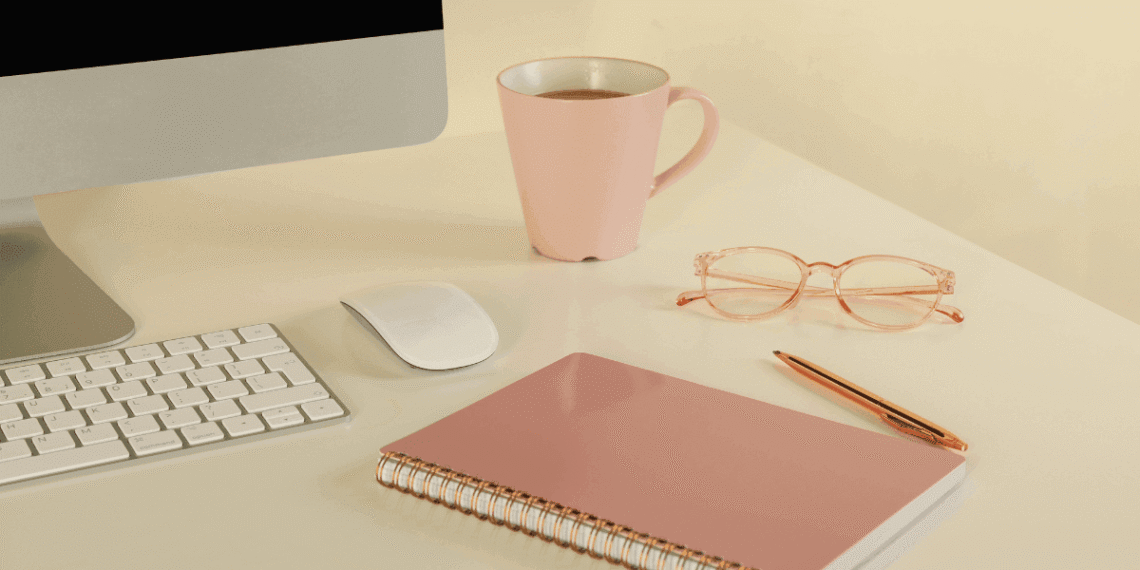 Escritorio de emprendedora con ordenador, taza rosa, gafas y cuaderno representando un espacio de trabajo organizado