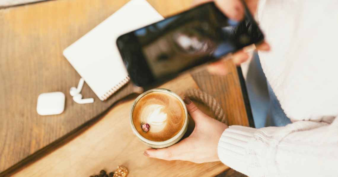 Mujer fotografiando un café con el móvil junto a un cuaderno y unos auriculares.