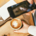 Mujer fotografiando un café con el móvil junto a un cuaderno y unos auriculares.